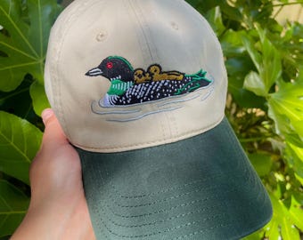 Può includere: Un cappellino da baseball beige con visiera verde scuro e un bottone verde scuro sulla parte superiore. La parte anteriore presenta un ricamo di una strolaga bianca e nera con un occhio rosso, che trasporta due piccoli pulcini marroni.