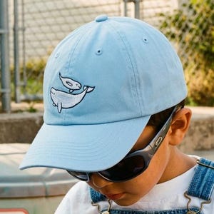 Könnte beinhalten: Eine hellblaue Baseballkappe mit einer gestickten Grafik von zwei weißen Walen auf der Vorderseite. Die Kappe hat einen gebogenen Schirm und eine strukturierte Krone, was für einen klassischen Look mit einem verspielten maritimen Motiv sorgt.