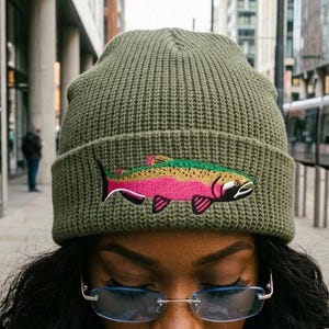 Puede incluir: Un gorro de punto acanalado color verde oliva con un gr&aacute;fico bordado de una trucha arco&iacute;ris con cuerpo rosa, verde y negro. El gorro tiene un dobladillo plegado y se lleva con gafas rectangulares sin montura con cristales azul claro.