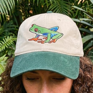 May include: A baseball cap with a beige crown and a dark green brim. The front of the cap features an embroidered graphic of a green tree frog with red eyes, blue accents on its legs, and orange feet. The cap has a structured front panel.