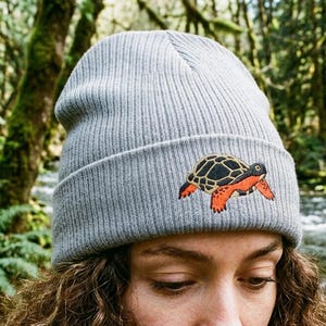 Può includere: Un berretto in maglia a coste grigio chiaro con un ricamo raffigurante una tartaruga con guscio nero e arti arancioni. Il cappello presenta un risvolto piegato ed è realizzato in un tessuto testurizzato con una dettagliata illustrazione.