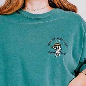 May include: Teal blue t-shirt with a graphic of a coffee cup and the text "Support Your Local Coffee Shop".