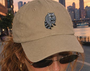 May include: A tan baseball cap featuring an embroidered graphic of a grey and black raccoon on the front panel. The cap has a structured crown, a curved brim, and a button at the top center. The embroidery depicts a raccoon in a standing pose.