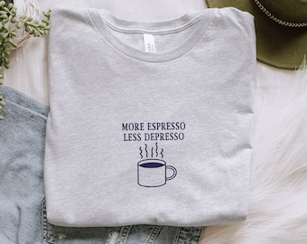 Puede incluir: Una camiseta gris con el texto "More Espresso Less Depresso" y una imagen de dibujos animados de una taza de café humeante.