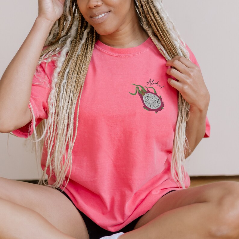 May include: A coral-colored t-shirt featuring a dragon fruit graphic with the word "Aloha!" in a script font. The dragon fruit is depicted with a purple exterior, green stems, and a white interior with black seeds. The shirt is a casual wear item.