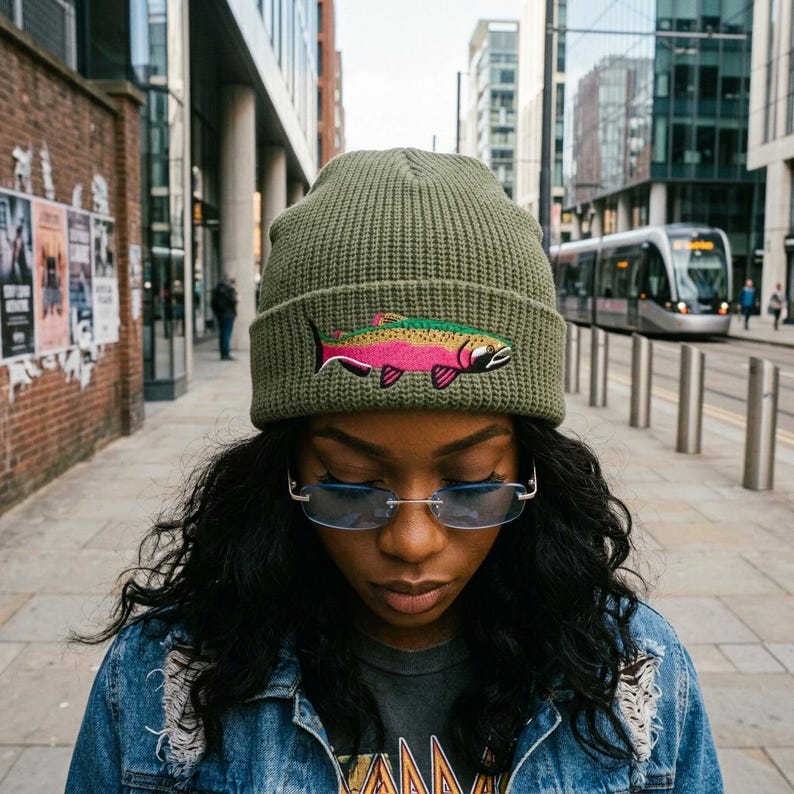 Puede incluir: Un gorro de punto acanalado color verde oliva con un gr&aacute;fico bordado de una trucha arco&iacute;ris con cuerpo rosa, verde y negro. El gorro tiene un dobladillo plegado y se lleva con gafas rectangulares sin montura con cristales azul claro.