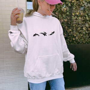 May include: A white hoodie with a black graphic of three orcas jumping. The hoodie has a hood and a kangaroo pocket.