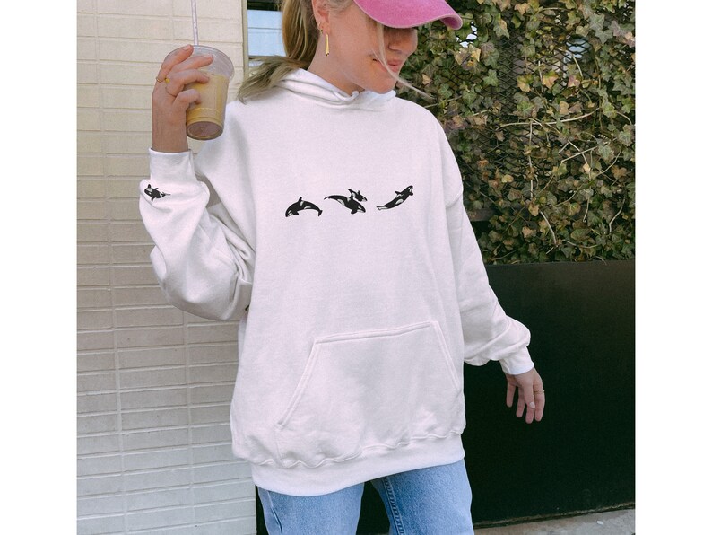 May include: A white hoodie with a black graphic of three orcas jumping. The hoodie has a hood and a kangaroo pocket.