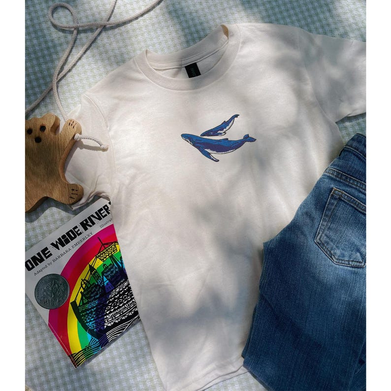 K&ouml;nnte beinhalten: Cremefarbenes T-Shirt mit einem blauen Wal-Design und einem kleineren Wal dar&uuml;ber. Das Shirt wird mit blauen Jeans gezeigt. Ein Holzspielzeug in B&auml;renform und ein Buch mit dem Titel "ONE WIDE RIVER" sind ebenfalls zu sehen.