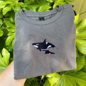K&ouml;nnte beinhalten: Ein graues T-Shirt mit einem gestickten Design von zwei Orcas, einem Erwachsenen und einem Jungtier. Die Orcas sind schwarz und wei&szlig;. Das Shirt ist gefaltet, das Design ist mittig auf der Vorderseite. Der Hintergrund ist gr&uuml;nes Laub.