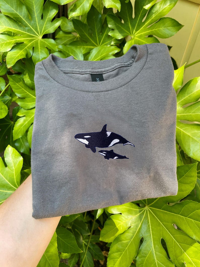 K&ouml;nnte beinhalten: Ein graues T-Shirt mit einem gestickten Design von zwei Orcas, einem Erwachsenen und einem Jungtier. Die Orcas sind schwarz und wei&szlig;. Das Shirt ist gefaltet, das Design ist mittig auf der Vorderseite. Der Hintergrund ist gr&uuml;nes Laub.