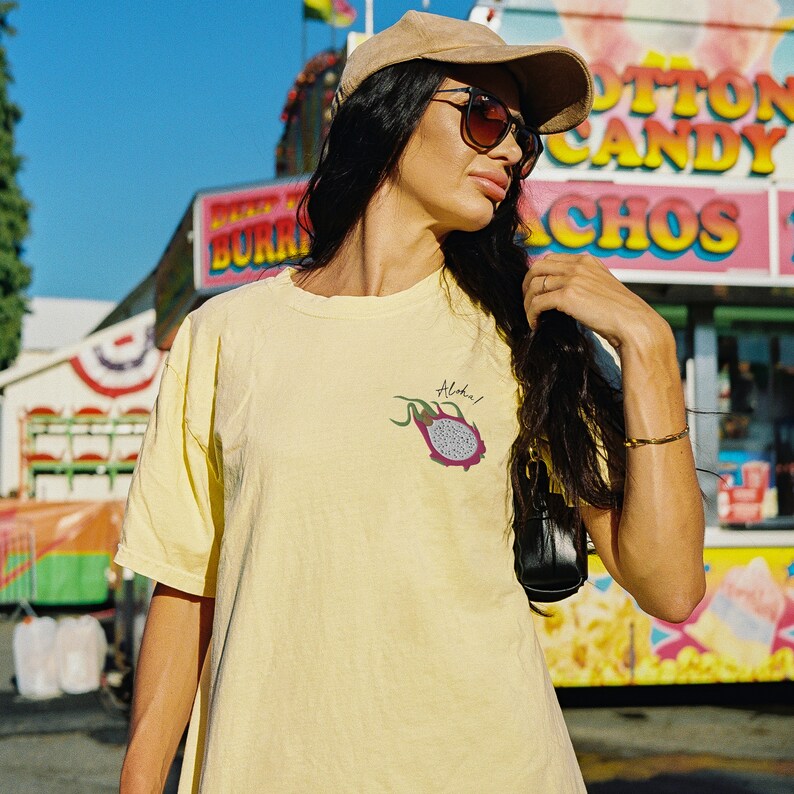 May include: A light yellow t-shirt with a small dragon fruit graphic and the word "Aloha!" printed on the front. The shirt is made of a soft material and is perfect for a casual day out. The background includes a fair with food vendors.