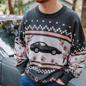 May include: A gray sweater featuring a car graphic, pink cherry blossoms, and geometric patterns. The sweater has a crew neck and long sleeves. The car is black and the flowers are pink. The sweater is worn with blue jeans.