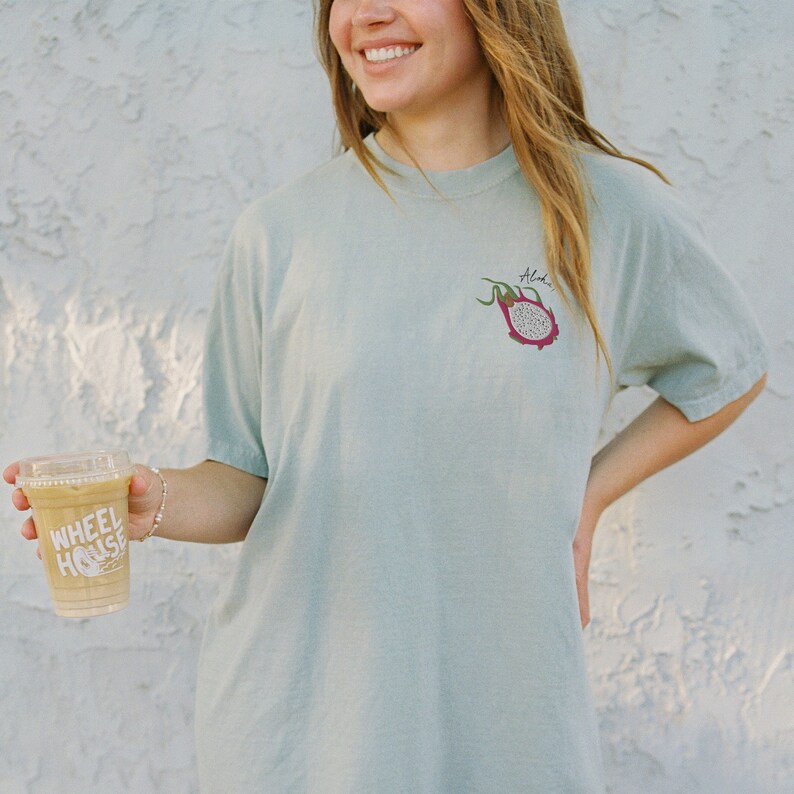 May include: A light green t-shirt with a small dragon fruit graphic and the word "Aloha" printed on the front. The shirt is paired with a clear plastic cup of coffee with the text "WHEEL HOUSE" printed on the side.
