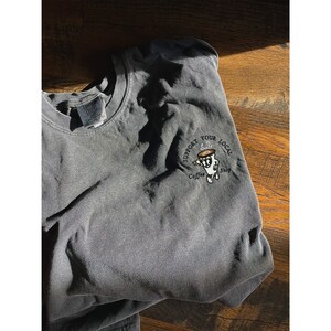 May include: A dark gray t-shirt with a white embroidered design that says "Support Your Local Coffee Shop" and features a cartoon coffee cup with steam coming out of it.