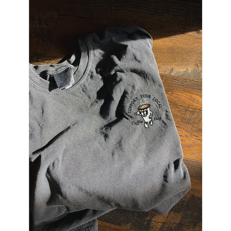 May include: A dark gray t-shirt with a white embroidered design that says "Support Your Local Coffee Shop" and features a cartoon coffee cup with steam coming out of it.
