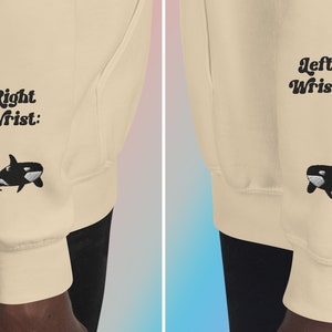 May include: A beige sweatshirt with black embroidered orcas on the sleeves. The left sleeve has the text "Left Wrist:" and the right sleeve has the text "Right Wrist:".