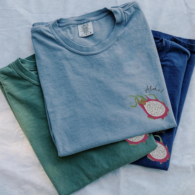 May include: Three folded t-shirts in shades of green, blue, and denim blue. Each shirt features a graphic of a dragon fruit with the word "Aloha" in script. The shirts are made of a soft material.