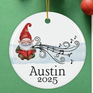 May include: A white ceramic Christmas ornament featuring a cartoon gnome wearing a red hat and glasses, holding a book with musical notes flowing from it. The ornament is personalized with the name "Austin" and the year "2025".