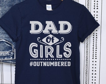 May include: A navy blue t-shirt with the text "DAD of GIRLS #OUTNUMBERED" in white. The shirt is hanging on a wooden hanger, with a pair of blue jeans on a black metal ladder in the background.