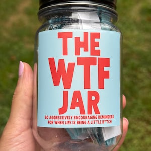 Op de afbeelding: Een doorzichtige plastic pot met een zwarte schroefdop en een lichtblauw etiket met rode tekst die luidt THE WTF JAR 60 AGGRESSIVELY ENCOURAGING REMINDERS FOR WHEN LIFE IS BEING A LITTLE B*TCH. De pot is gevuld met kleine gevouwen briefjes.