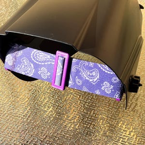 May include: A black welding helmet with a purple paisley patterned head strap. The strap has a white paisley design and a bright purple adjustment buckle. The helmet is designed for eye protection during welding.