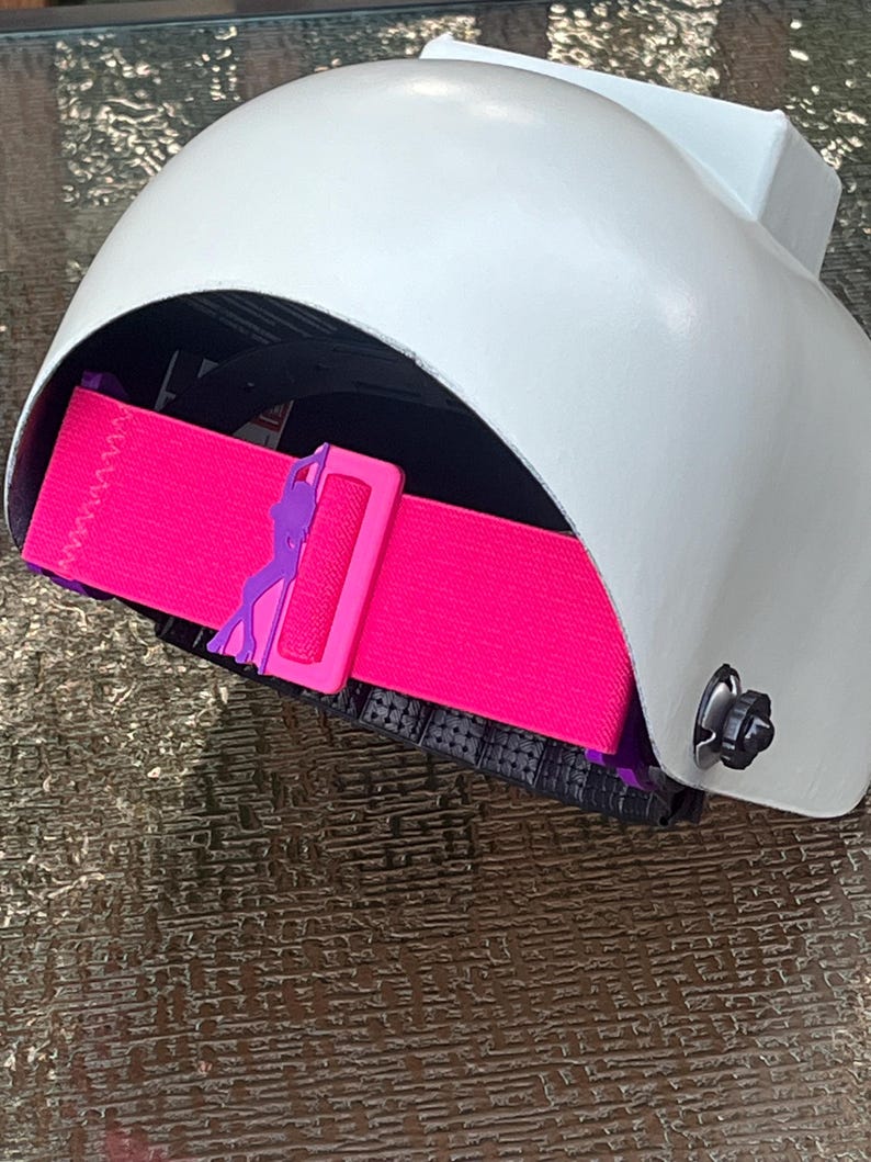 May include: A white helmet with a bright pink strap and a purple buckle. The helmet has a curved design and a black interior. The strap is adjustable and features a decorative element on the buckle.