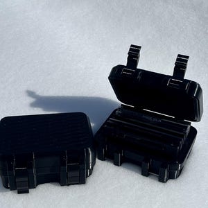 May include: Two black, rectangular cases with hinged lids. One case is closed, and the other is open, revealing several dark, rectangular objects inside. The cases have a textured surface and secure latches. The cases are on a white surface.