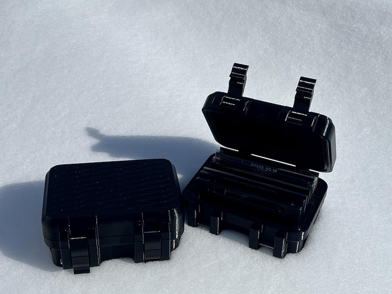 May include: Two black, rectangular cases with hinged lids. One case is closed, and the other is open, revealing several dark, rectangular objects inside. The cases have a textured surface and secure latches. The cases are on a white surface.
