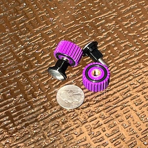 May include: Close-up of three purple and black metal objects, possibly fasteners or knobs, with a silver coin for scale. The objects have a ribbed design and a shiny finish. The background is a textured brown surface.