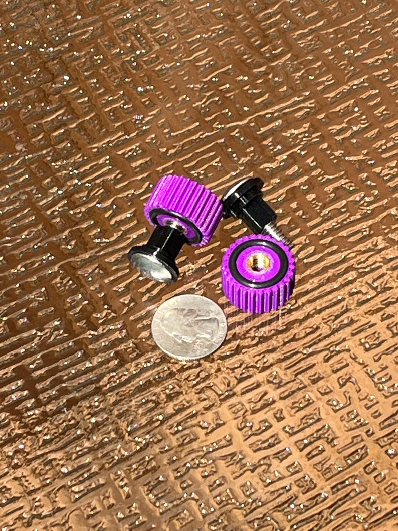 May include: Close-up of three purple and black metal objects, possibly fasteners or knobs, with a silver coin for scale. The objects have a ribbed design and a shiny finish. The background is a textured brown surface.