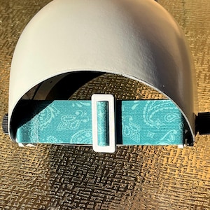 May include: A white welding helmet featuring a teal paisley patterned strap and a white buckle. The helmet is constructed from a smooth, light-coloured material, designed to shield the wearer's face and eyes during welding.