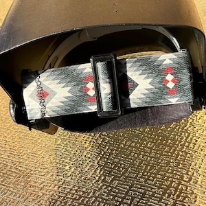 May include: A welding helmet with a patterned strap. The strap features a geometric design in shades of grey, white, and red. The helmet is dark in colour and has a curved shape. The strap is secured with a black plastic buckle.