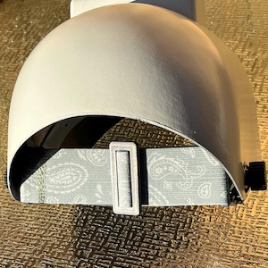 May include: A white welding helmet with a curved visor and a paisley patterned head strap. The strap is adjustable with a silver-coloured buckle. The helmet has a black adjustment knob on the side.