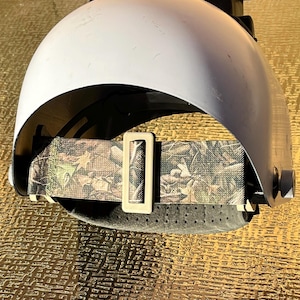 May include: A white welding helmet with a camouflage head strap and a beige buckle. The helmet has a curved design and a dark interior. The strap is made of a fabric with a green, brown, and beige pattern.