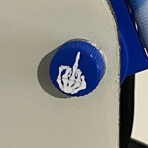 Puede incluir: Primer plano de una tapa de botella azul con un gesto de mano blanco y estilizado, un dedo medio. La tapa est&aacute; fijada a una superficie blanca.