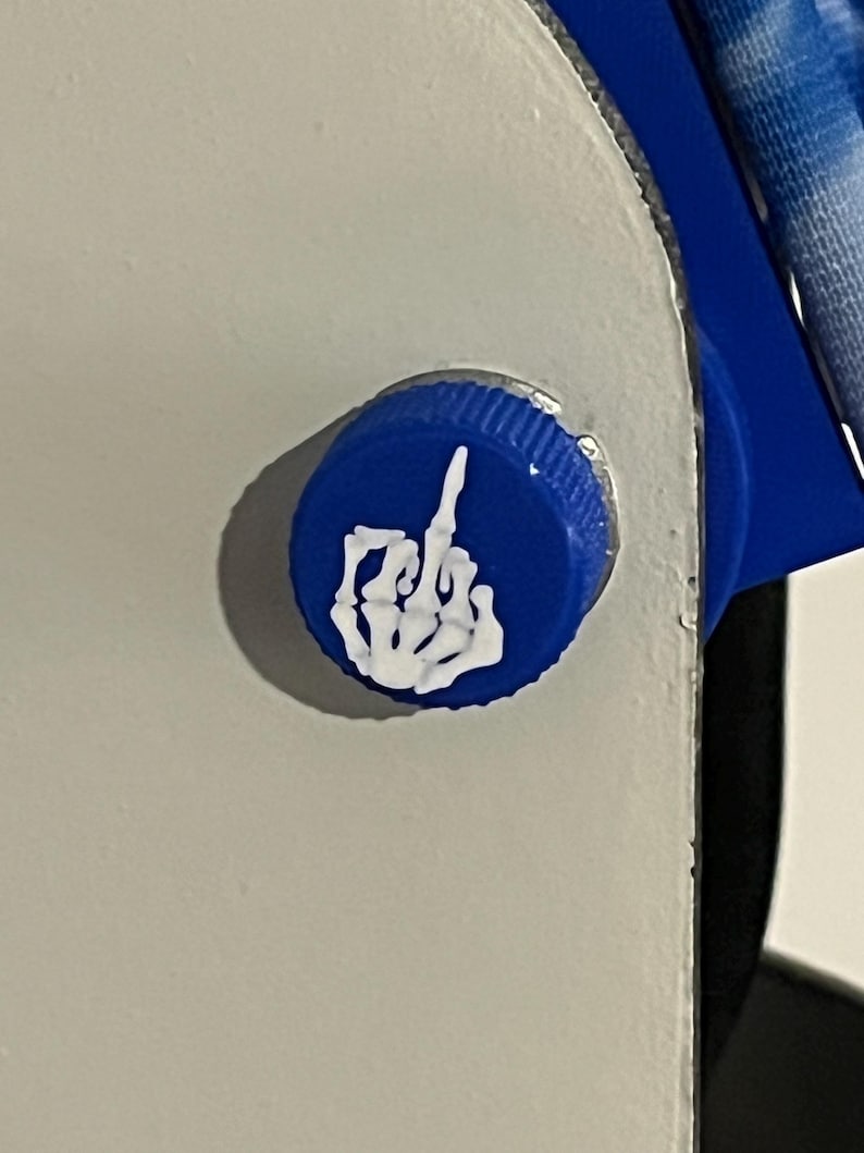Puede incluir: Primer plano de una tapa de botella azul con un gesto de mano blanco y estilizado, un dedo medio. La tapa est&aacute; fijada a una superficie blanca.
