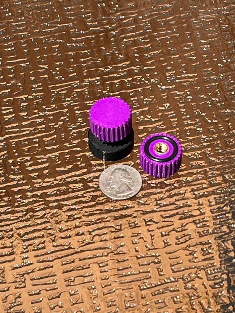 Puede incluir: Tres peque&ntilde;as piezas mec&aacute;nicas moradas y negras se muestran junto a una moneda de veinticinco centavos estadounidense. Las piezas tienen un dise&ntilde;o estriado y una forma circular. La moneda es plateada y muestra un perfil de George Washington.