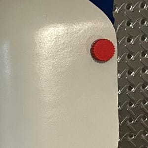 Puede incluir: Primer plano de un objeto blanco con un bot&oacute;n rojo y circular en el lateral. Se ve una tela azul en la parte superior. El objeto tiene una superficie lisa y brillante. El fondo es una superficie met&aacute;lica texturizada.
