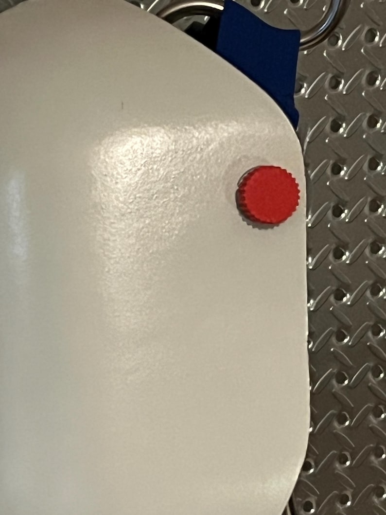 Puede incluir: Primer plano de un objeto blanco con un bot&oacute;n rojo y circular en el lateral. Se ve una tela azul en la parte superior. El objeto tiene una superficie lisa y brillante. El fondo es una superficie met&aacute;lica texturizada.