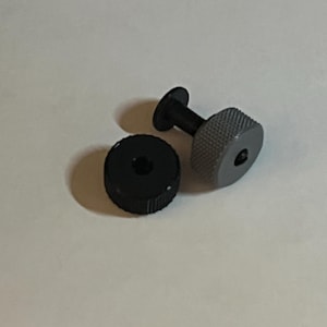 May include: A black and grey plastic screw with a threaded head and a round base. The screw is designed for use with a small electronic device.