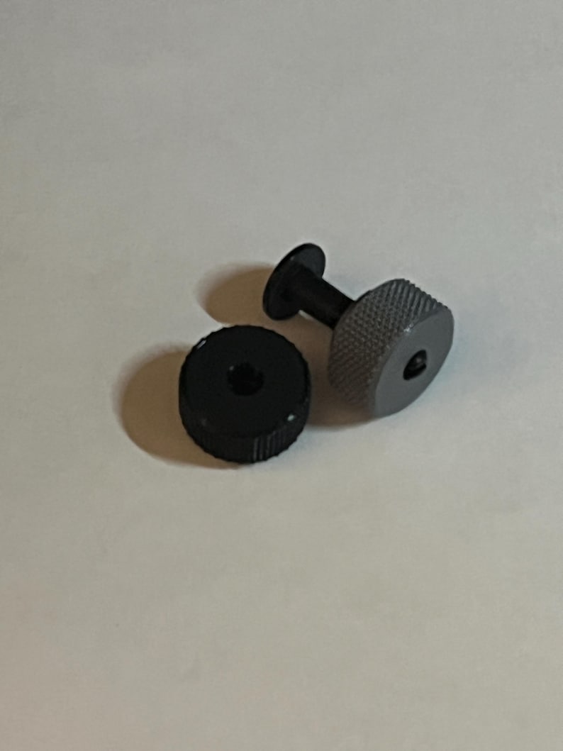 May include: A black and grey plastic screw with a threaded head and a round base. The screw is designed for use with a small electronic device.