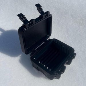 May include: An open, black, rectangular case with a hinged lid and two latches. The interior of the case has several vertical dividers. The case is on a white, snowy surface.