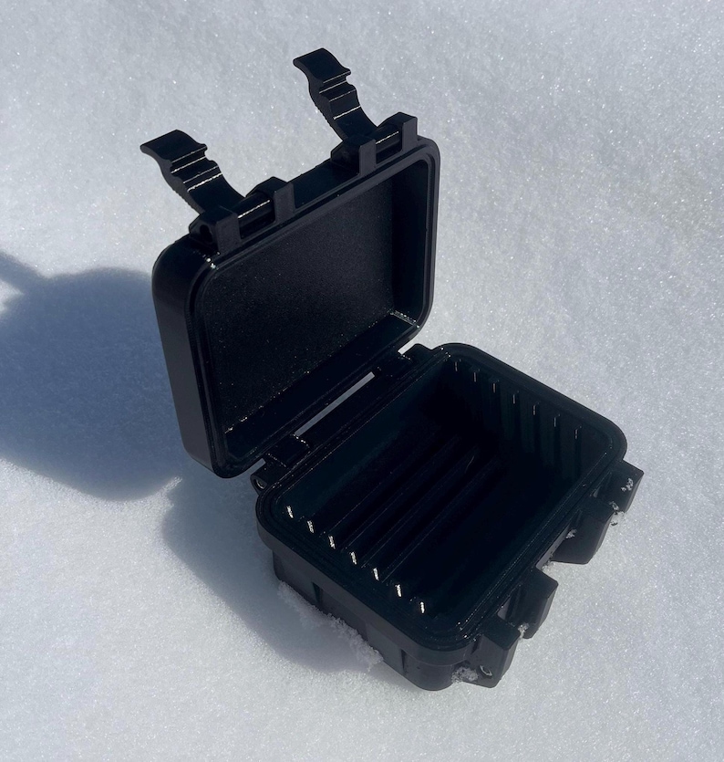 May include: An open, black, rectangular case with a hinged lid and two latches. The interior of the case has several vertical dividers. The case is on a white, snowy surface.