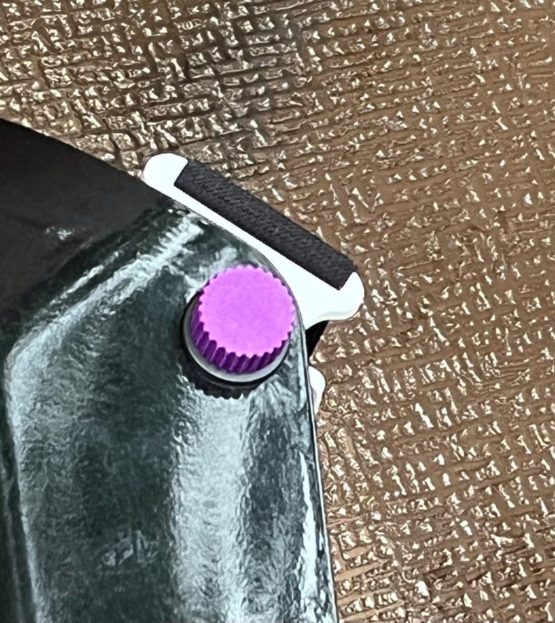 May include: Close-up of a black object with a shiny, reflective surface. A small, round, purple button with a ridged edge is visible. A black strap and white trim are also present.