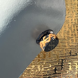 May include: A black and gold high-heeled shoe with a dollar sign design on the sole. The shoe has a black upper and a gold accent around the heel. The background is a textured gold surface.
