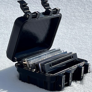 May include: A black, open, rectangular case with a hinged lid, containing several rectangular slides. The case has a textured surface and secure latches. The slides are labeled with text. The case is on a white surface.