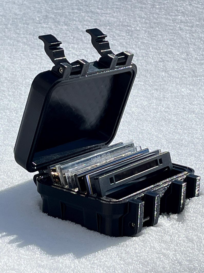May include: A black, open, rectangular case with a hinged lid, containing several rectangular slides. The case has a textured surface and secure latches. The slides are labeled with text. The case is on a white surface.