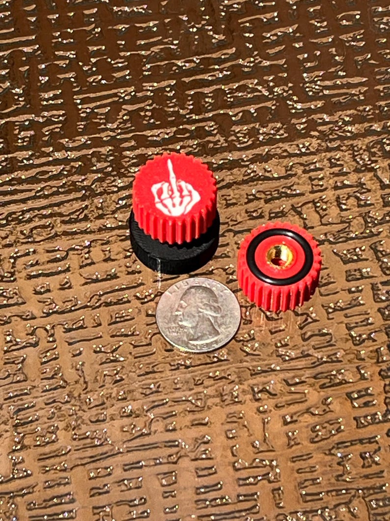 Puede incluir: Dos perillas decorativas rojas y negras con un gr&aacute;fico de dedo medio y una moneda de veinticinco centavos. Las perillas tienen un borde estriado y un gr&aacute;fico blanco. La moneda es plateada y muestra un perfil.