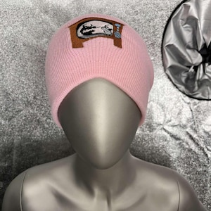 Puede incluir: Gorro de punto rosa con una imagen bordada en marrón y blanco de un televisor con un personaje de dibujos animados mirándolo.